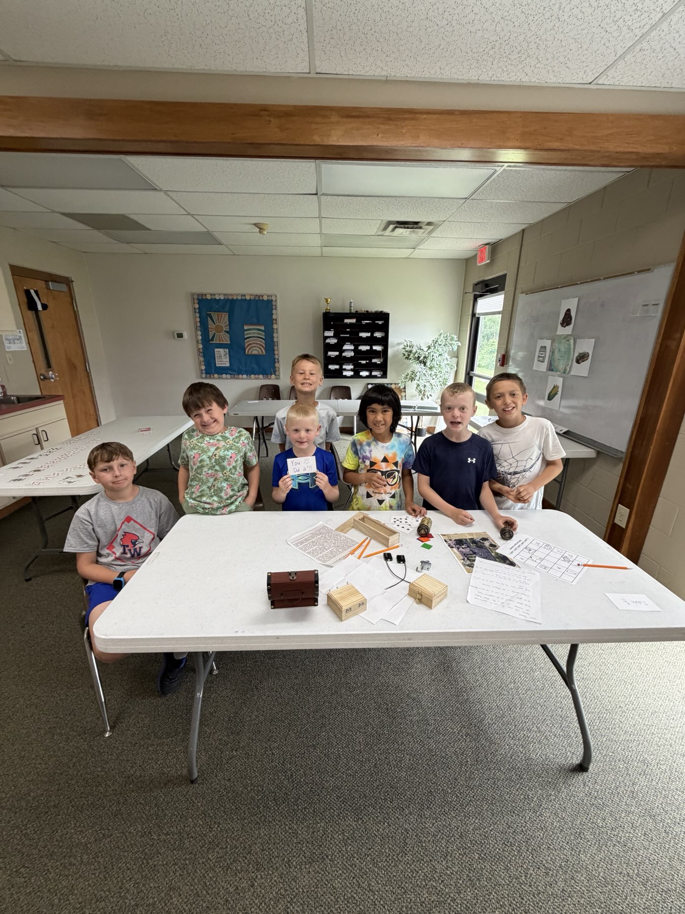 Seven campers around a table of puzzle boxes and clue sheets