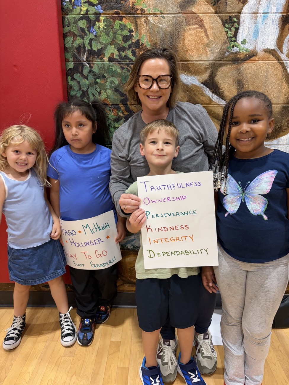 A teacher and students holding a poster that lists Truthfulness, Ownership, Perseverance, Kindness, Integrity, and Dependability